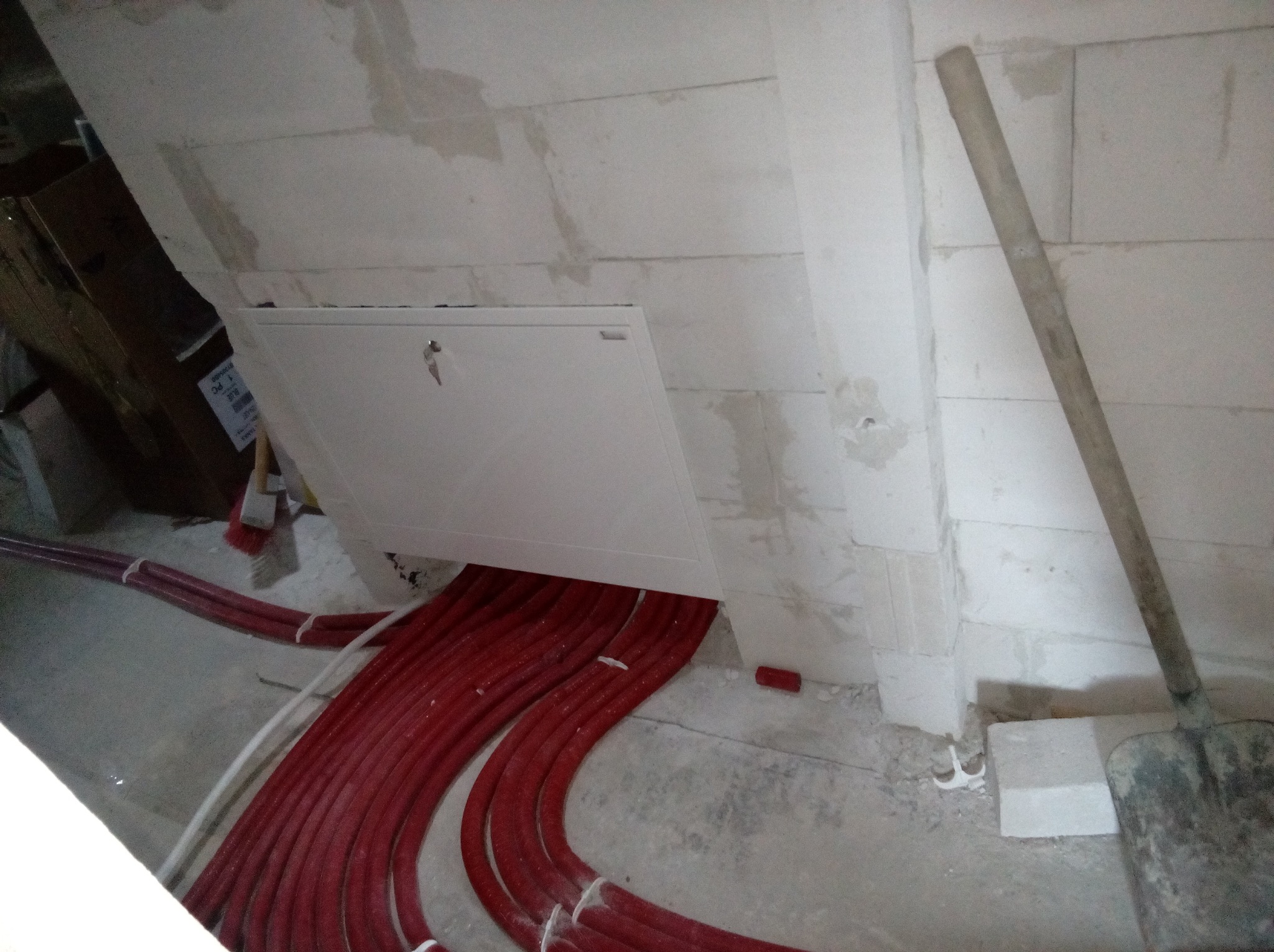 Czerwone rury instalacyjne wychodzące spod białej klapy rewizyjnej w ścianie z betonu komórkowego, obok stoi brudna łopata, widoczne fragmenty innych materiałów budowlanych.
