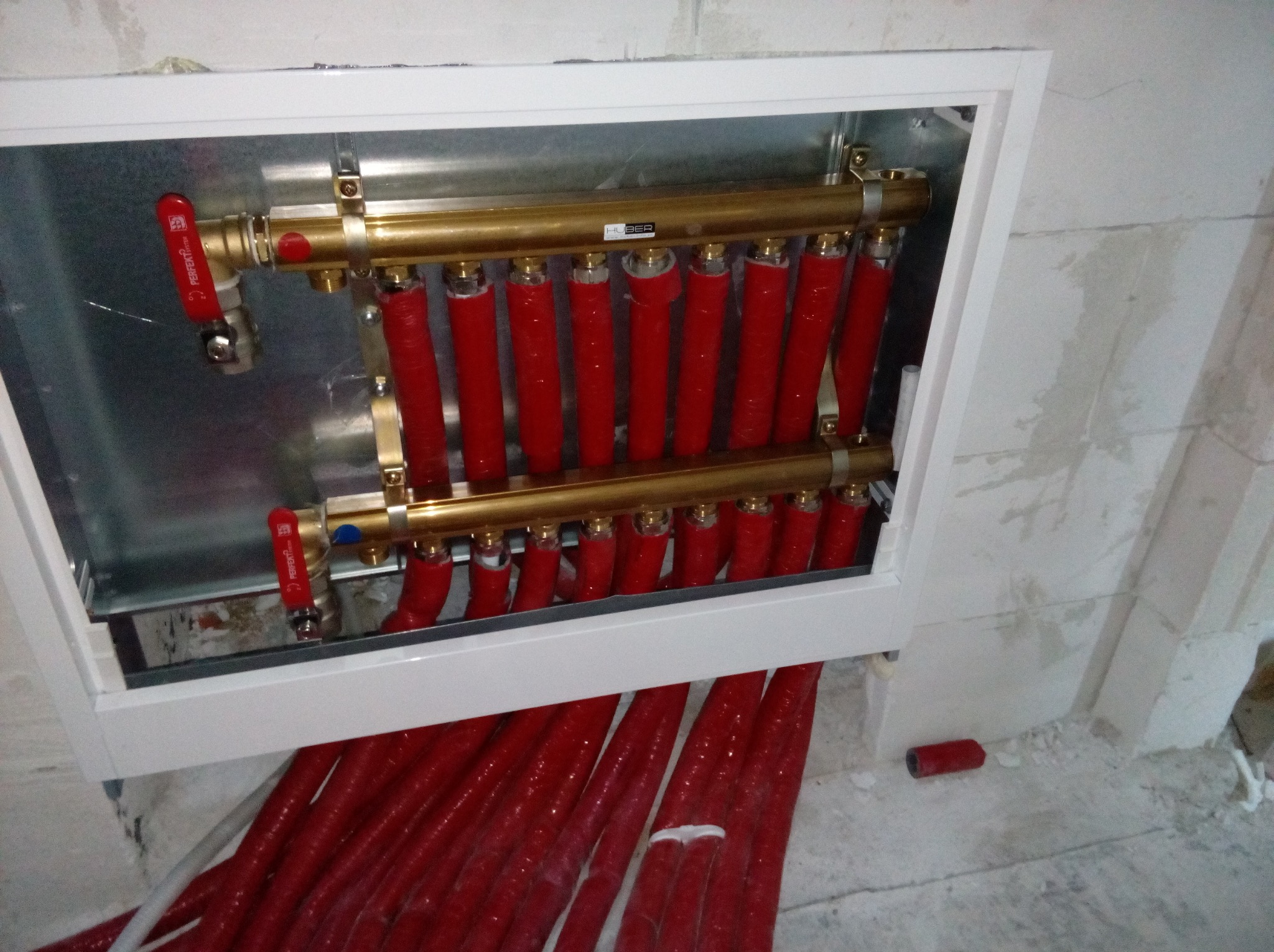 Rozdzielacz ogrzewania podłogowego z czerwonymi rurami w białej skrzynce montażowej, widok z góry na surową ścianę.