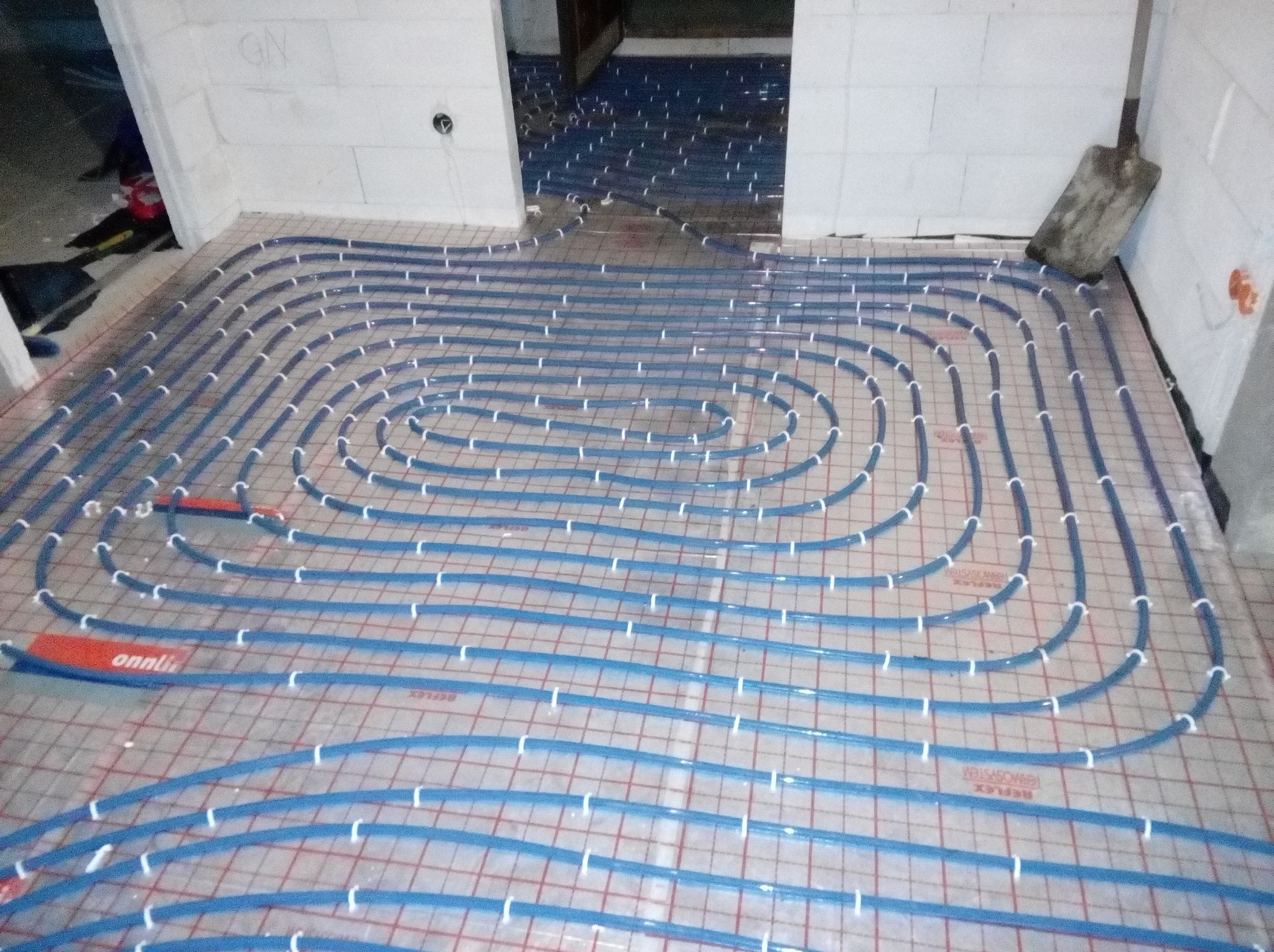 Niebieskie rury ogrzewania podłogowego, mocowane do siatki na podłodze, widok z góry, fragment łopaty oparty o ścianę z bloczków.