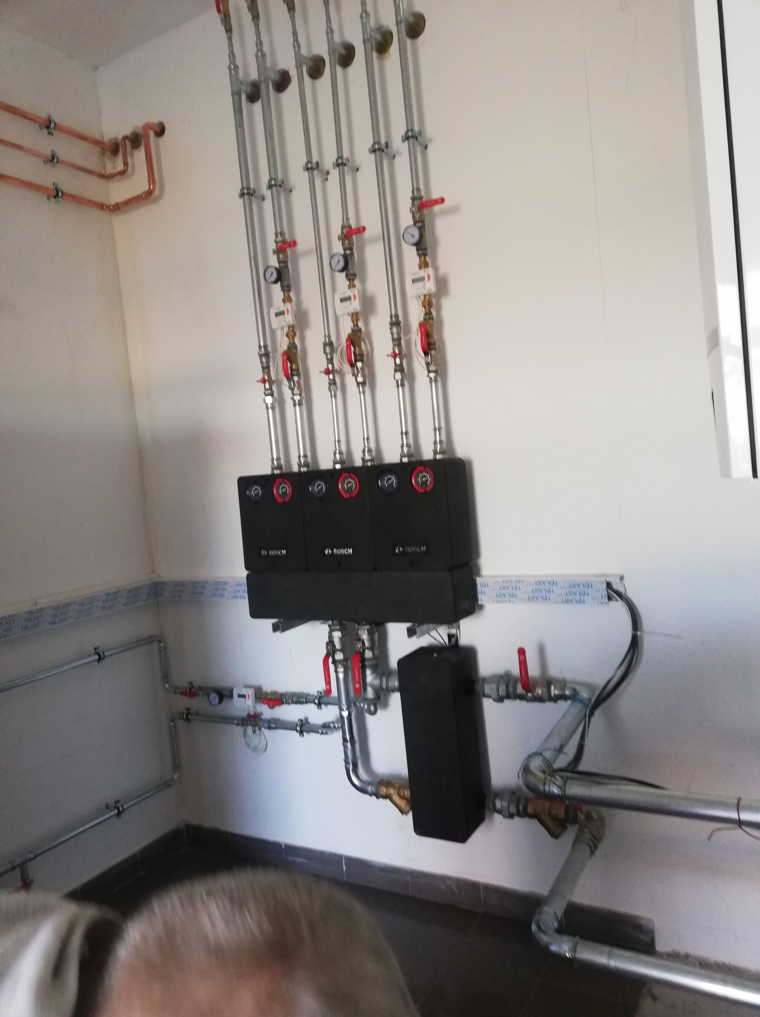 Rozdzielacz ogrzewania z czterema obiegami, z czarnymi modułami Bosch, manometrami i zaworami, instalacja wykonana w pomieszczeniu z płytkami na ścianach i podłodze.