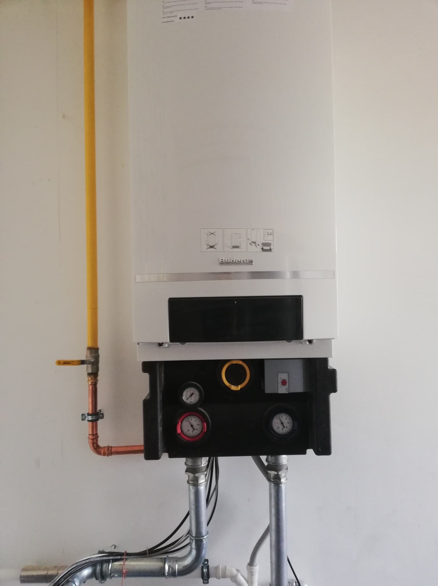 Nowoczesny, wiszący piec gazowy marki Buderus, z widocznymi manometrami, rurami miedzianymi i żółtym przewodem gazowym, zamontowany na białej ścianie.