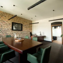 Sala konferencyjna z dużym drewnianym stołem, zielonymi krzesłami, ceglaną ścianą i nowoczesnym oświetleniem w biurze.