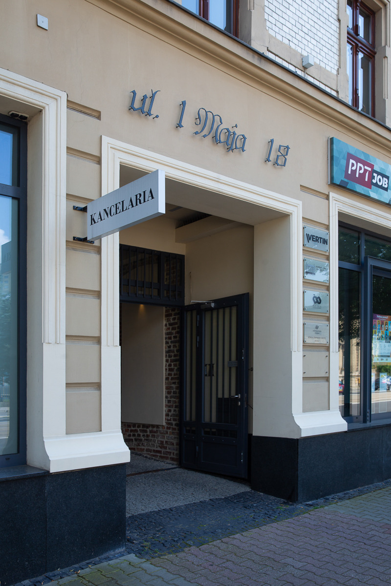 Elewacja budynku z szyldem 'Kancelaria' i adresem 'ul. 1 Maja 18', otwarte drzwi wejściowe.