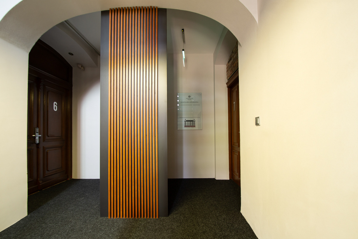 Wnętrze kancelarii prawnej: korytarz z drewnianymi panelami, drzwiami oznaczonymi numerami 6 i 7 oraz tabliczką informacyjną na ścianie.