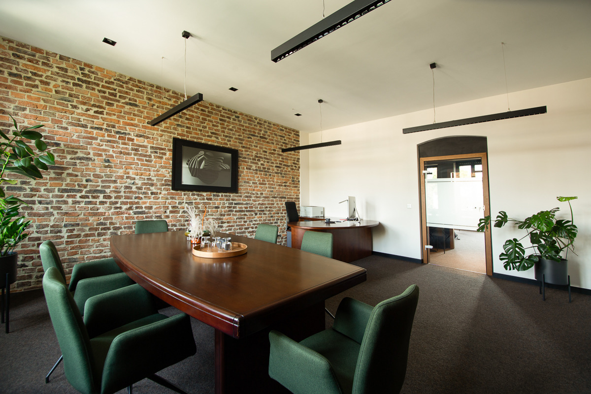 Sala konferencyjna z dużym drewnianym stołem, zielonymi krzesłami, ceglaną ścianą i nowoczesnym oświetleniem w biurze.