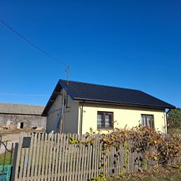 DachyPiotrowski - Żółty dom jednorodzinny z nowym, czarnym dachem z dachówki, widoczny zza drewnianego płotu z winoroślą, na tle błękitnego nieba.