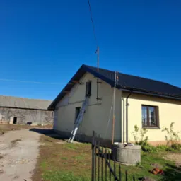 Dom jednorodzinny z nowym, czarnym dachem z blachodachówki, drabina oparta o ścianę, widoczna stara zabudowa gospodarcza w tle, błękitne niebo.