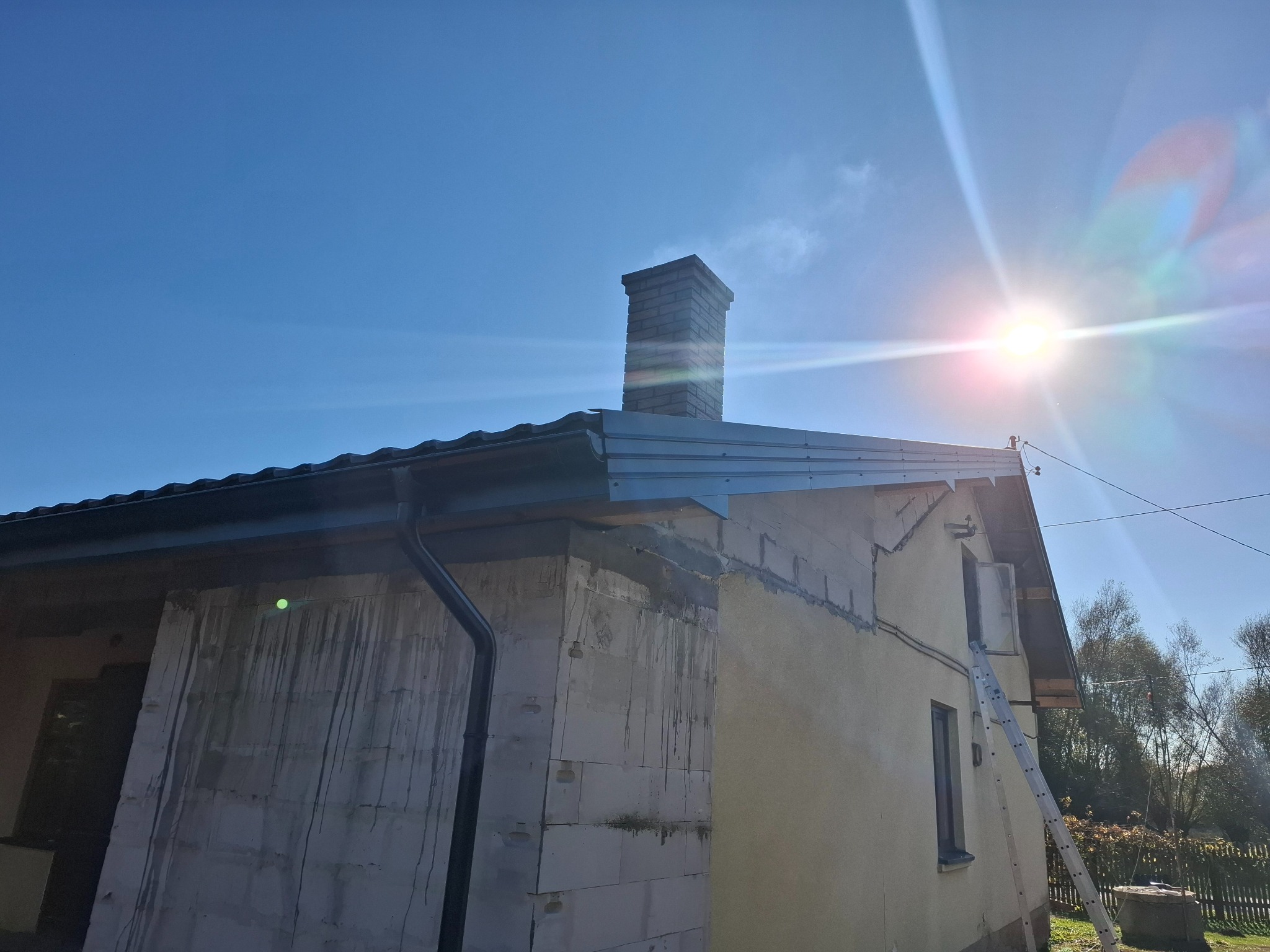 Dom jednorodzinny w trakcie remontu elewacji i dachu, widoczna nowa blacha na dachu, rynna, komin z cegły i ściany częściowo ocieplone, z drabina opartą o ścianę.