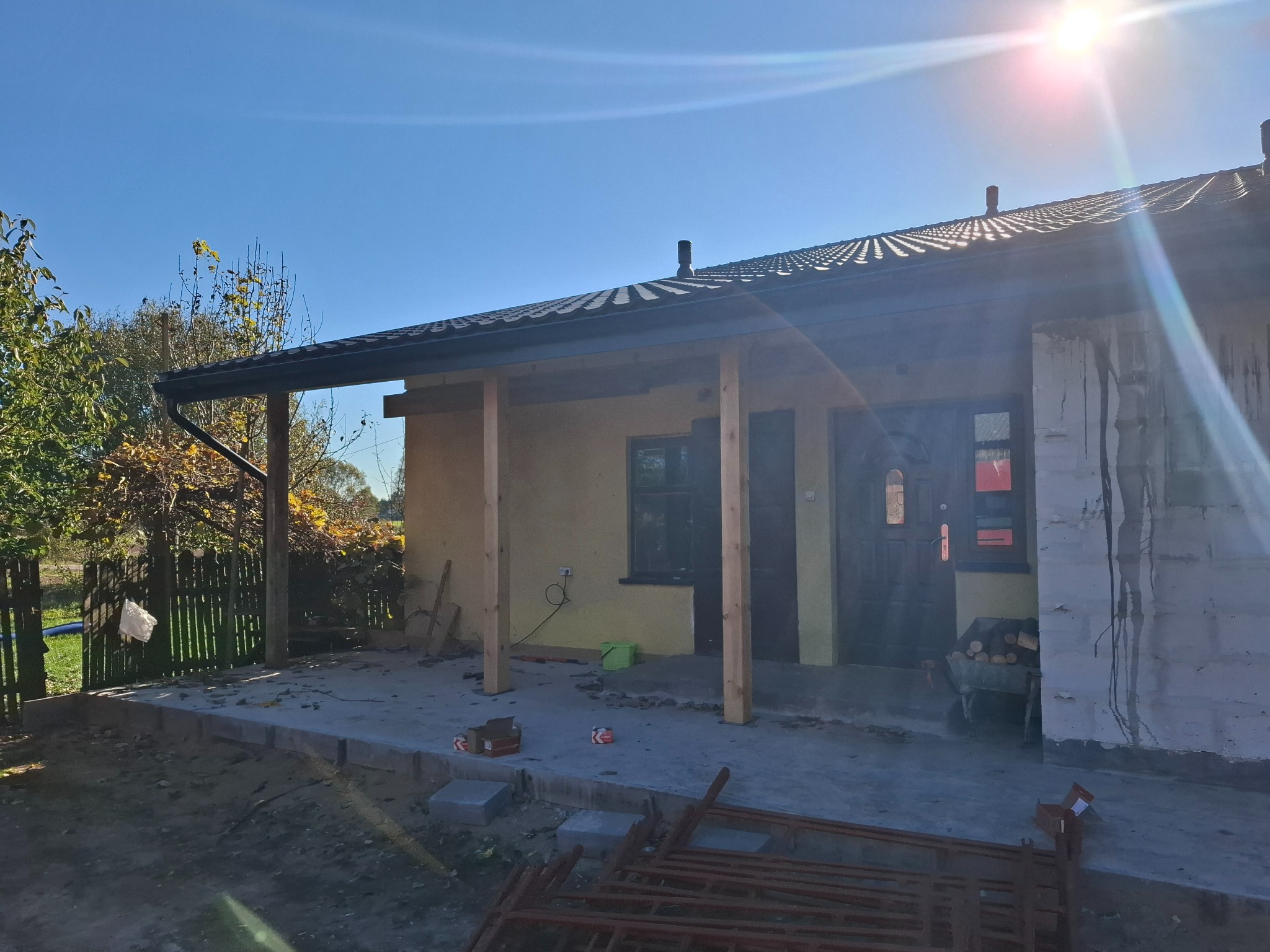 Dom w trakcie remontu z widocznym nowym dachem, drewnianymi słupami podtrzymującymi zadaszenie werandy i przygotowanym betonowym podłożem, elementy rusztowania na pierwszym planie.