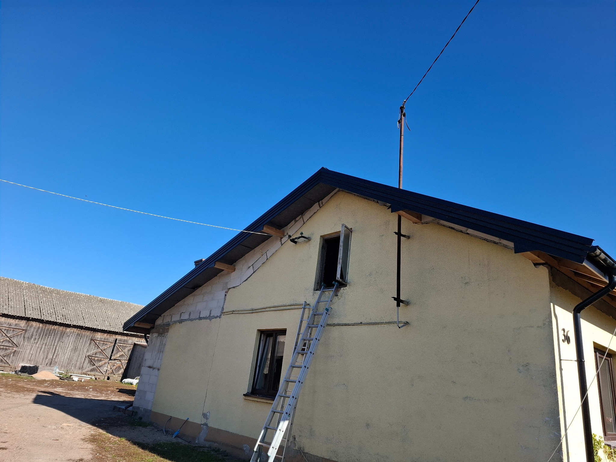Dom jednorodzinny w trakcie remontu elewacji z widoczną drabiną opartą o okno na poddaszu, fragment dachu z czarną blachodachówką i budynek gospodarczy w tle.