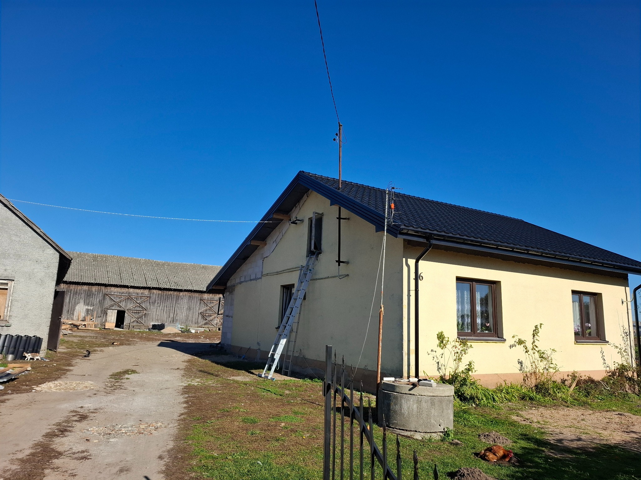 Dom jednorodzinny z nowym, czarnym dachem z blachodachówki, drabina oparta o ścianę, widoczna stara zabudowa gospodarcza w tle, błękitne niebo.