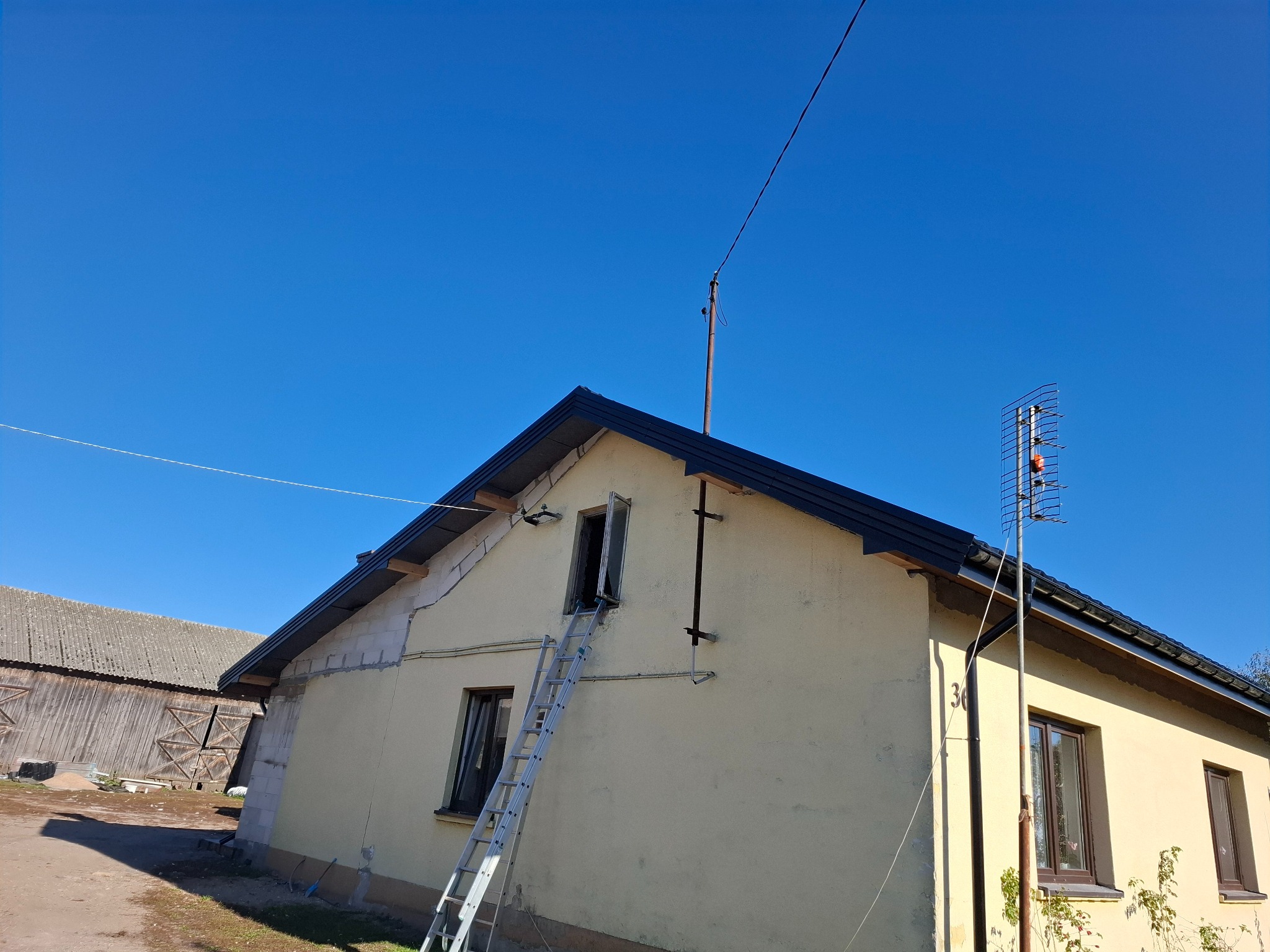 Elewacja domu jednorodzinnego w trakcie remontu, widoczna drabina przystawiona do okna na poddaszu, świeżo położone obróbki blacharskie dachu, w tle budynek gospodarczy.