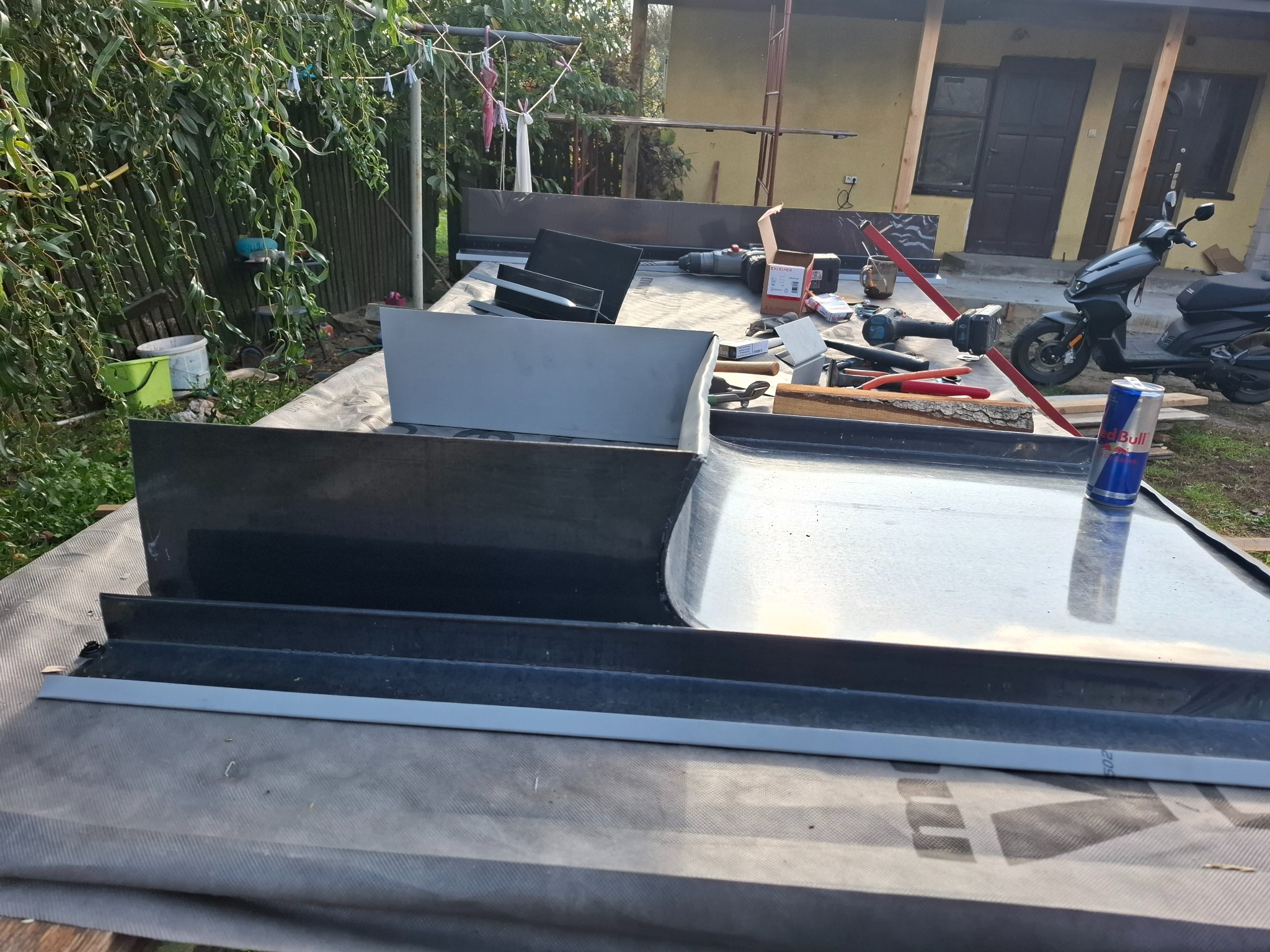 Przygotowanie elementów blacharskich na dachu w Płońsku, widoczne narzędzia, wkrętarka, puszka napoju energetycznego, elementy rynny i obróbek blacharskich na tle budynku i skutera.