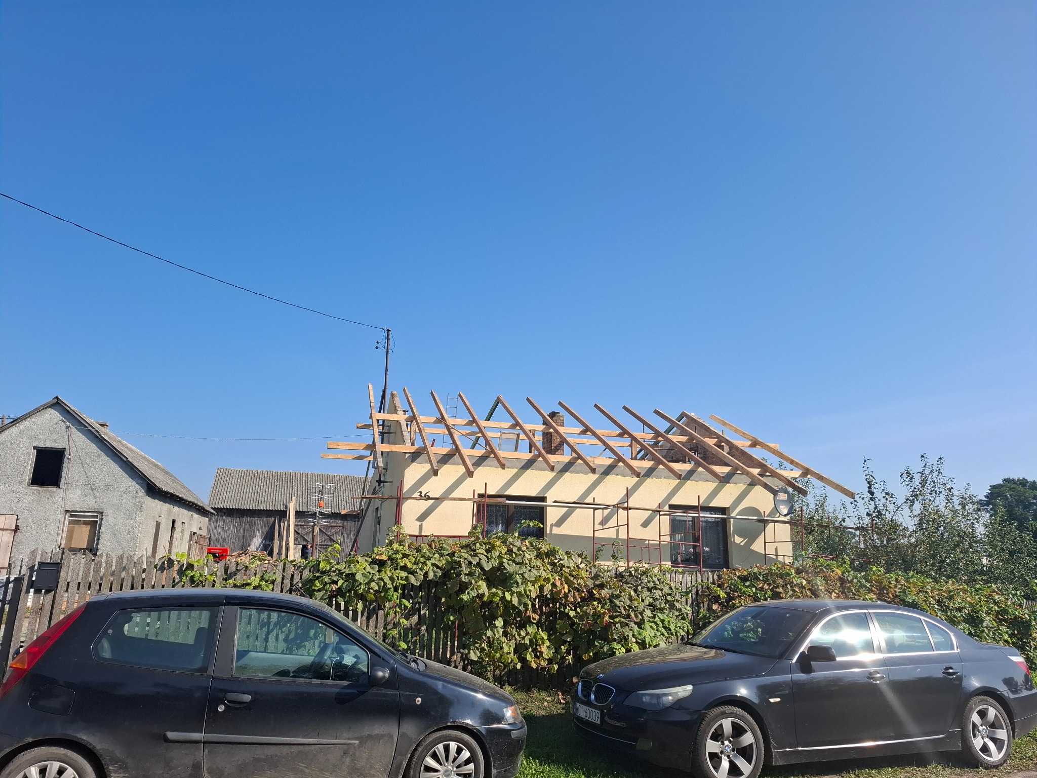 Konstrukcja drewniana dachu w trakcie budowy, widoczna zza ogrodzenia porośniętego zielenią, z zaparkowanymi dwoma samochodami na pierwszym planie i budynkami gospodarczymi w tle, w słoneczny dzień.