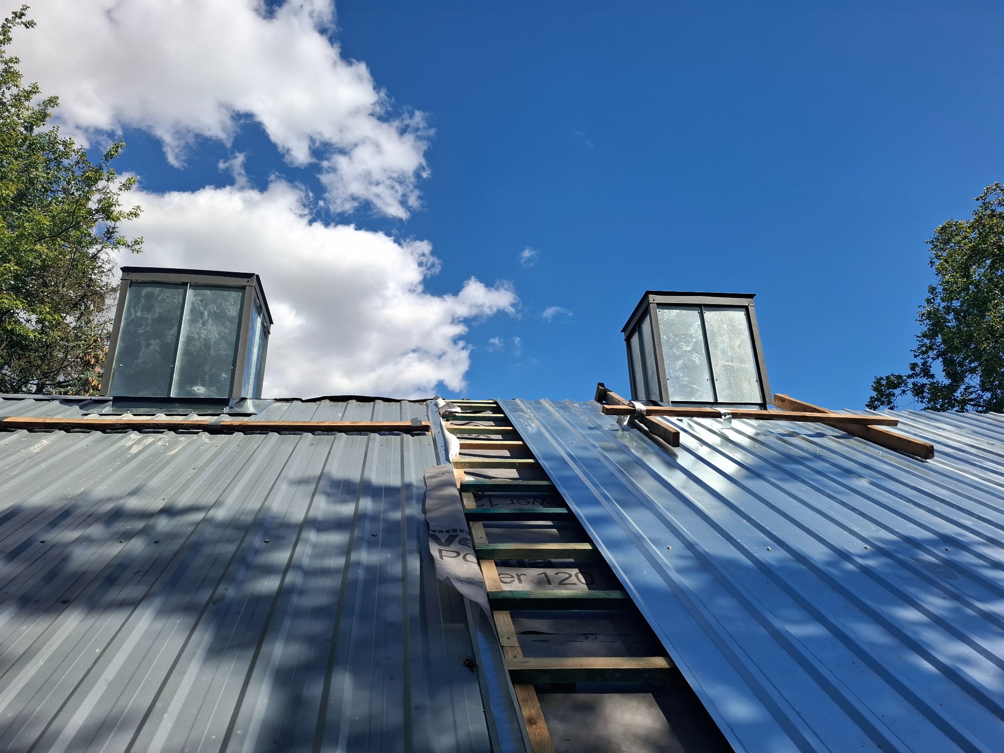 Metalowy dach z dwoma kominami w trakcie montażu, widoczne elementy konstrukcyjne, membrana i niebo z chmurami.