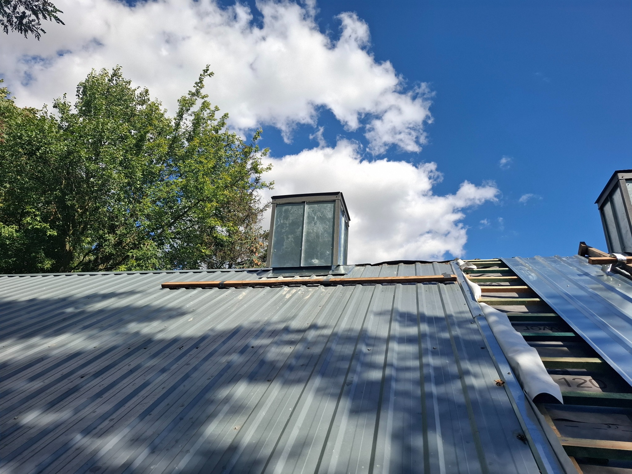 Szary blaszany dach w trakcie montażu, widoczne drewniane łaty, membrana dachowa, komin, fragment błękitnego nieba z chmurami i drzewo w tle.