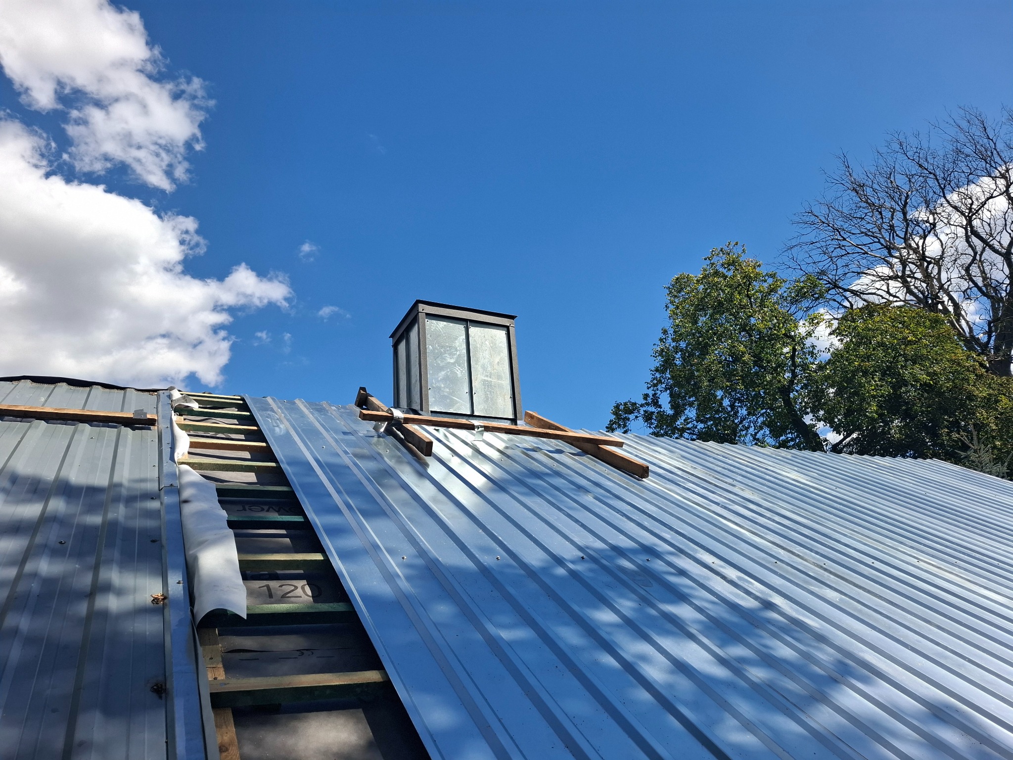 Szary dach z blachy trapezowej w trakcie montażu, widoczne elementy konstrukcji drewnianej, komin wentylacyjny i błękitne niebo z chmurami.