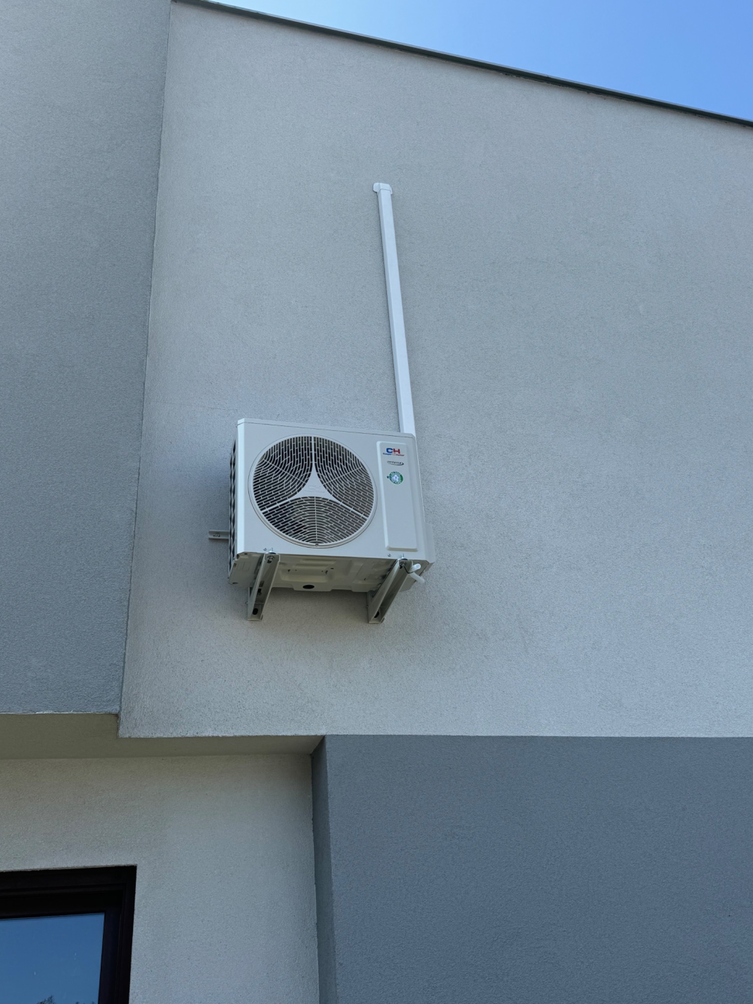 Biała jednostka zewnętrzna klimatyzacji marki CH zamontowana na szarej, teksturowanej ścianie budynku z widoczną rurą odprowadzającą skropliny.