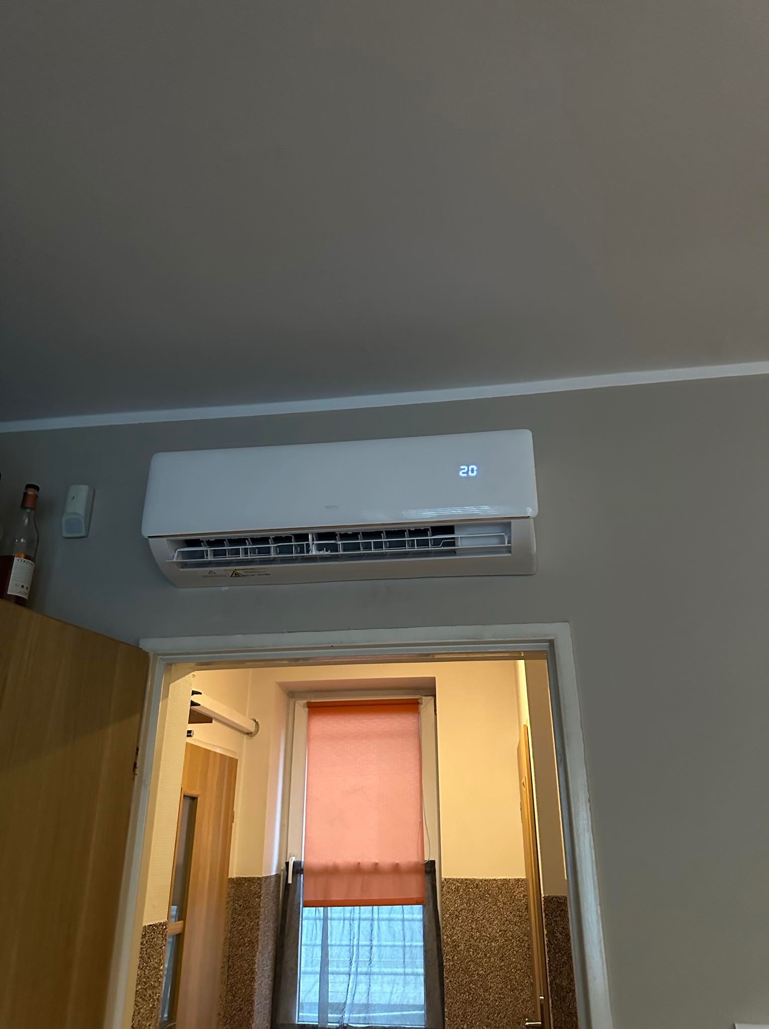 Biały klimatyzator ścienny z wyświetlaczem wskazującym temperaturę 20 stopni Celsjusza, zamontowany nad futryną drzwiową w szarym pomieszczeniu.