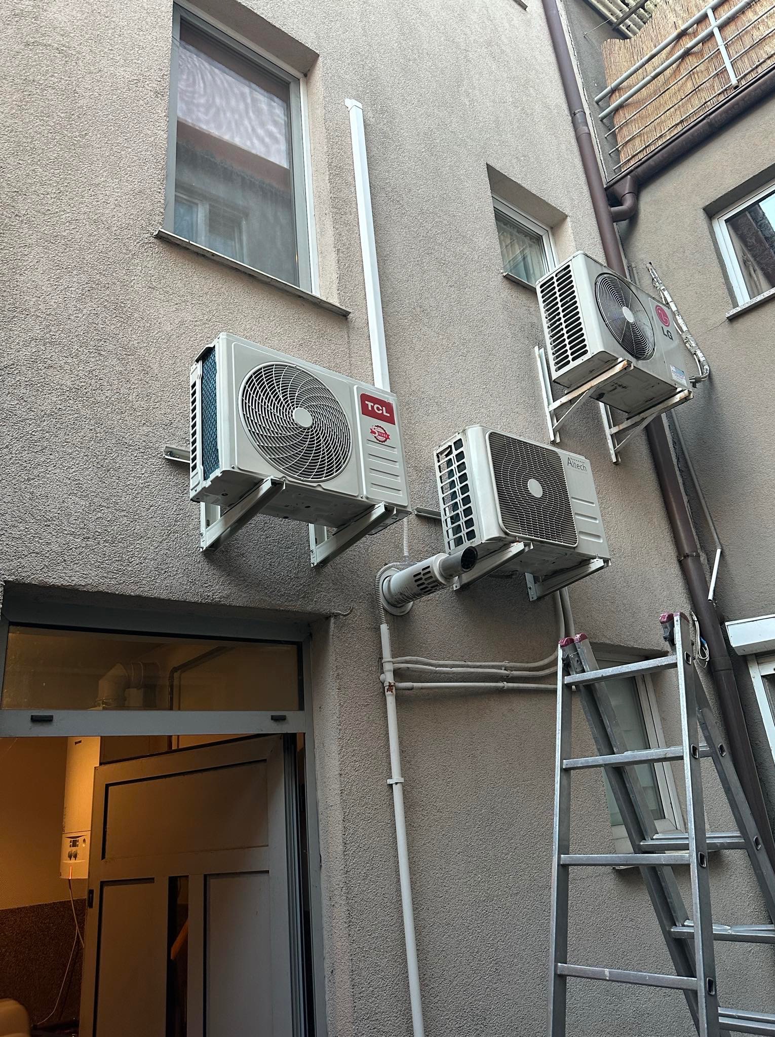 Trzy klimatyzatory różnych marek (TCL, Altech, LG) zamontowane na elewacji budynku, widoczna drabina oparta o ścianę.