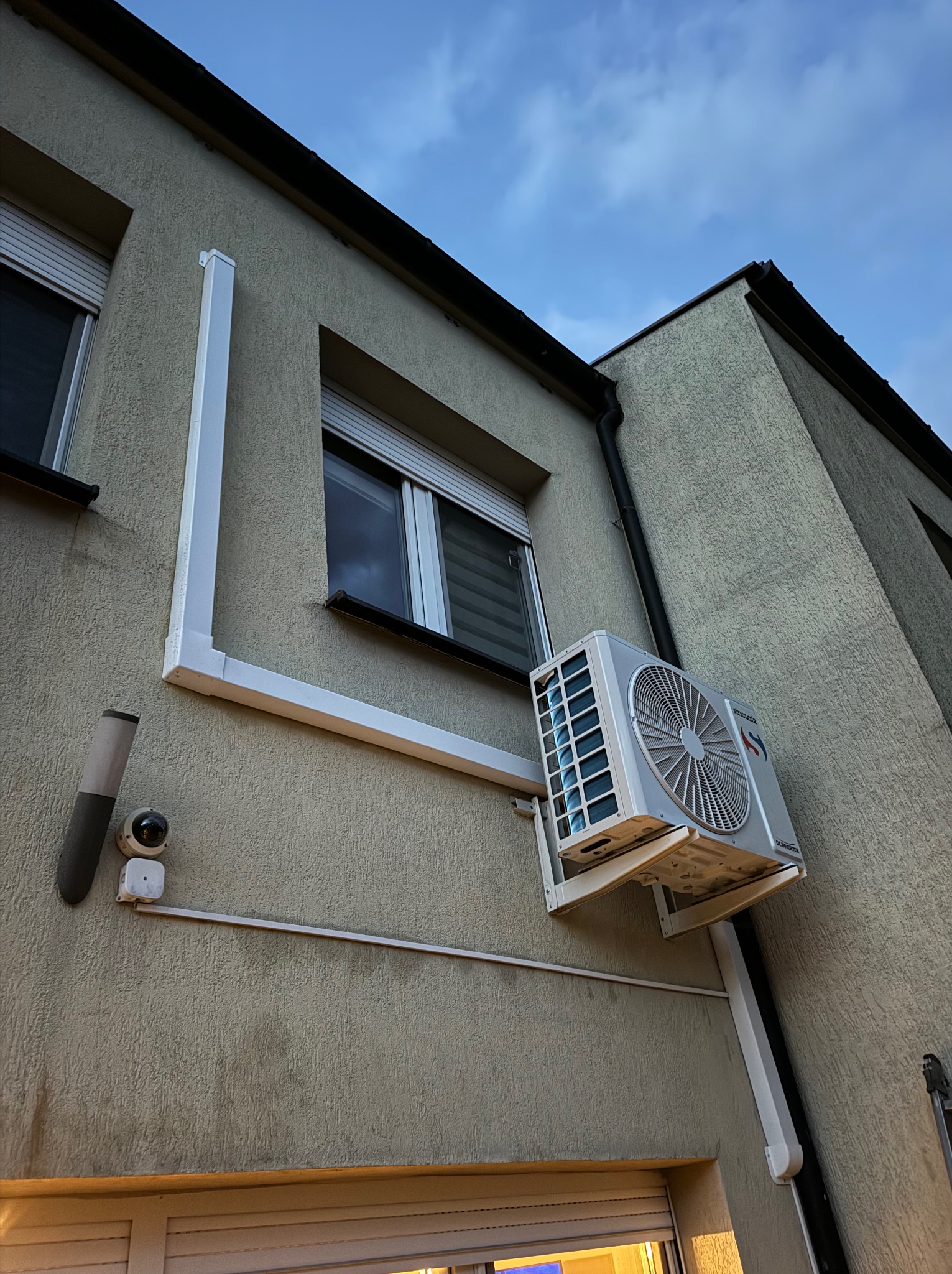 Zewnętrzna jednostka klimatyzacji zamontowana na elewacji budynku, widoczna instalacja odprowadzająca skropliny, ujęcie z dołu.