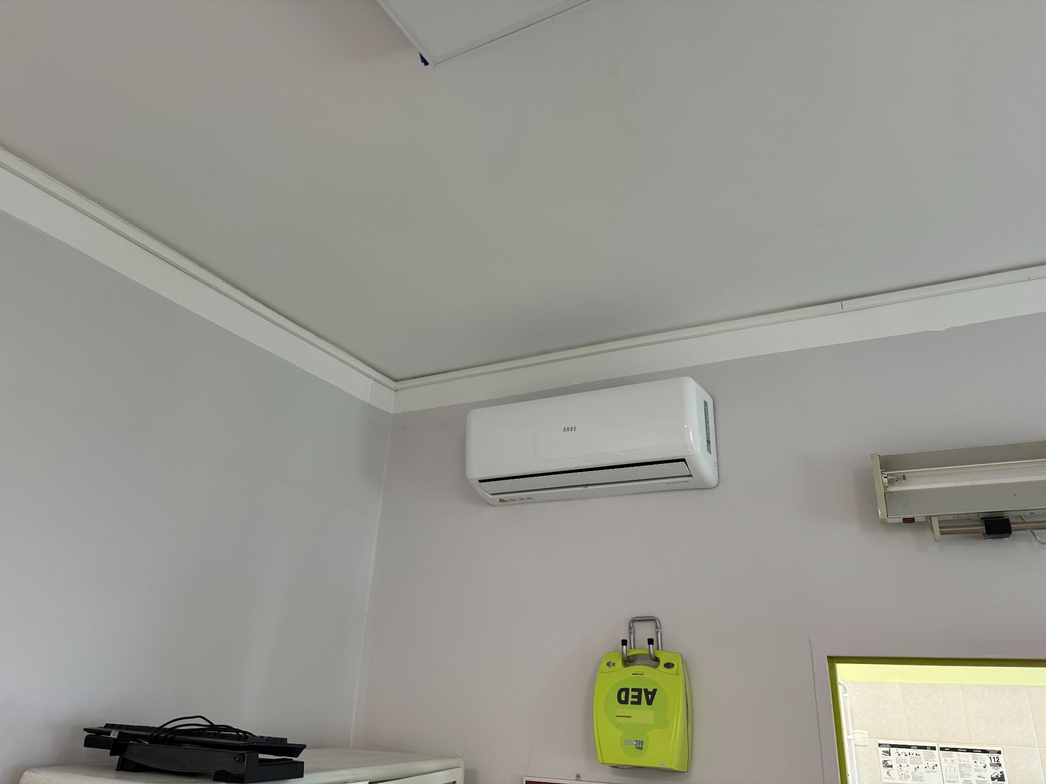 Biały klimatyzator ścienny marki ANDE zamontowany w pomieszczeniu z szarą ścianą, obok lampy fluorescencyjnej i defibrylatora AED.
