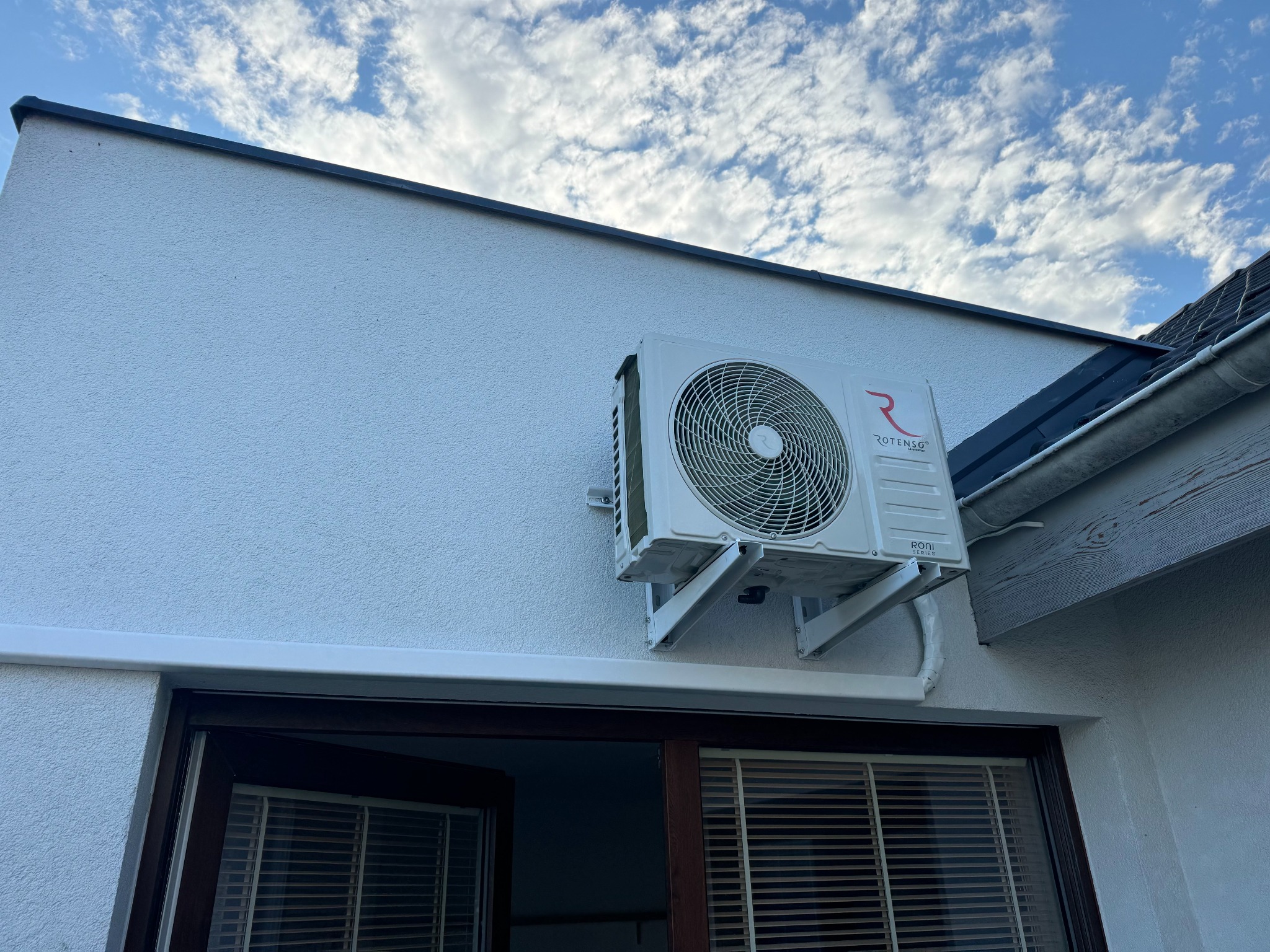 Biała jednostka zewnętrzna klimatyzacji marki Rotenso zamontowana na elewacji domu z widocznymi oknami i fragmentem dachu na tle błękitnego nieba z chmurami.