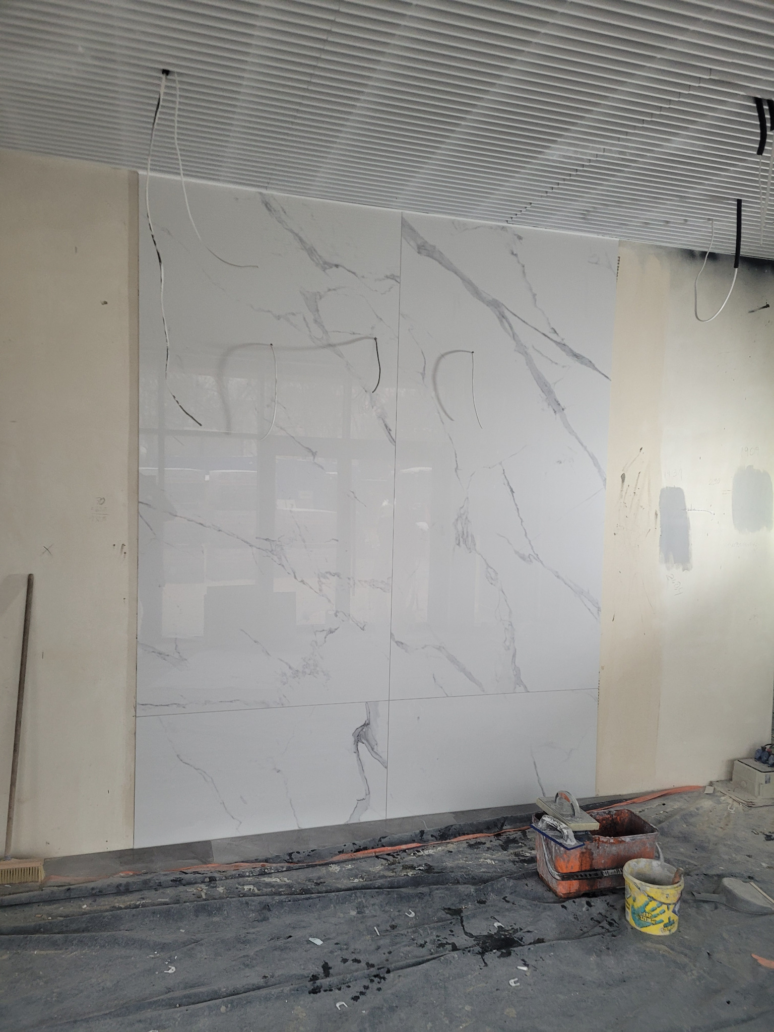 Ściana wyłożona wielkoformatowymi płytkami imitującymi marmur, widoczne przygotowania do montażu, narzędzia pracy i nieukończone kable elektryczne wystające z sufitu.