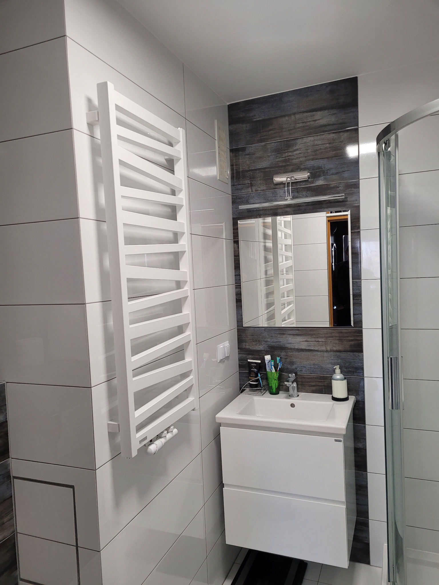 Nowoczesna łazienka z białymi płytkami, grzejnikiem drabinkowym, szafką z umywalką i fragmentem kabiny prysznicowej, akcentowana ciemną okładziną ściany za lustrem.