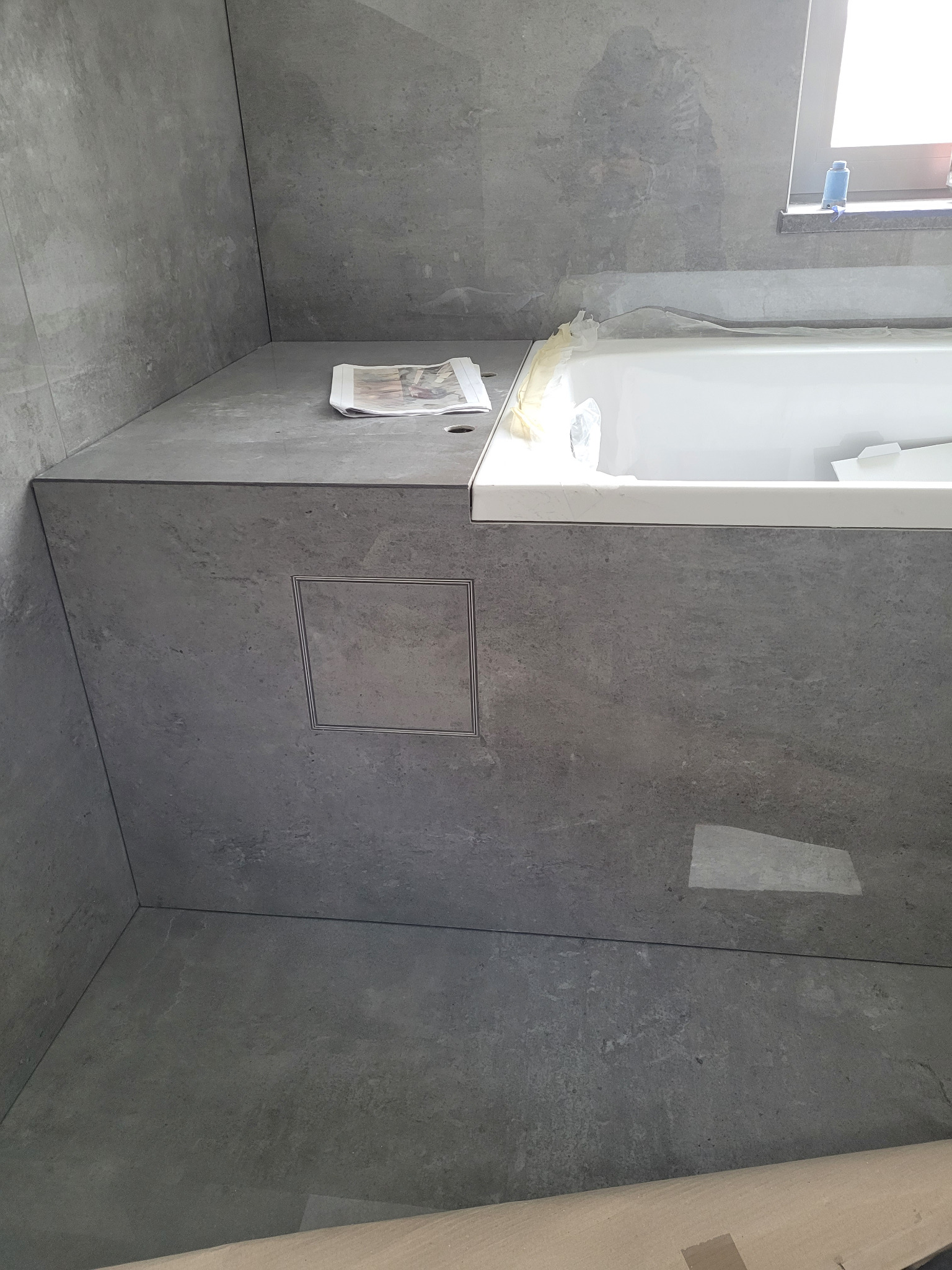 Szara łazienka z wanną obłożoną płytkami, widoczny odpływ liniowy w zabudowie i gazeta na blacie.