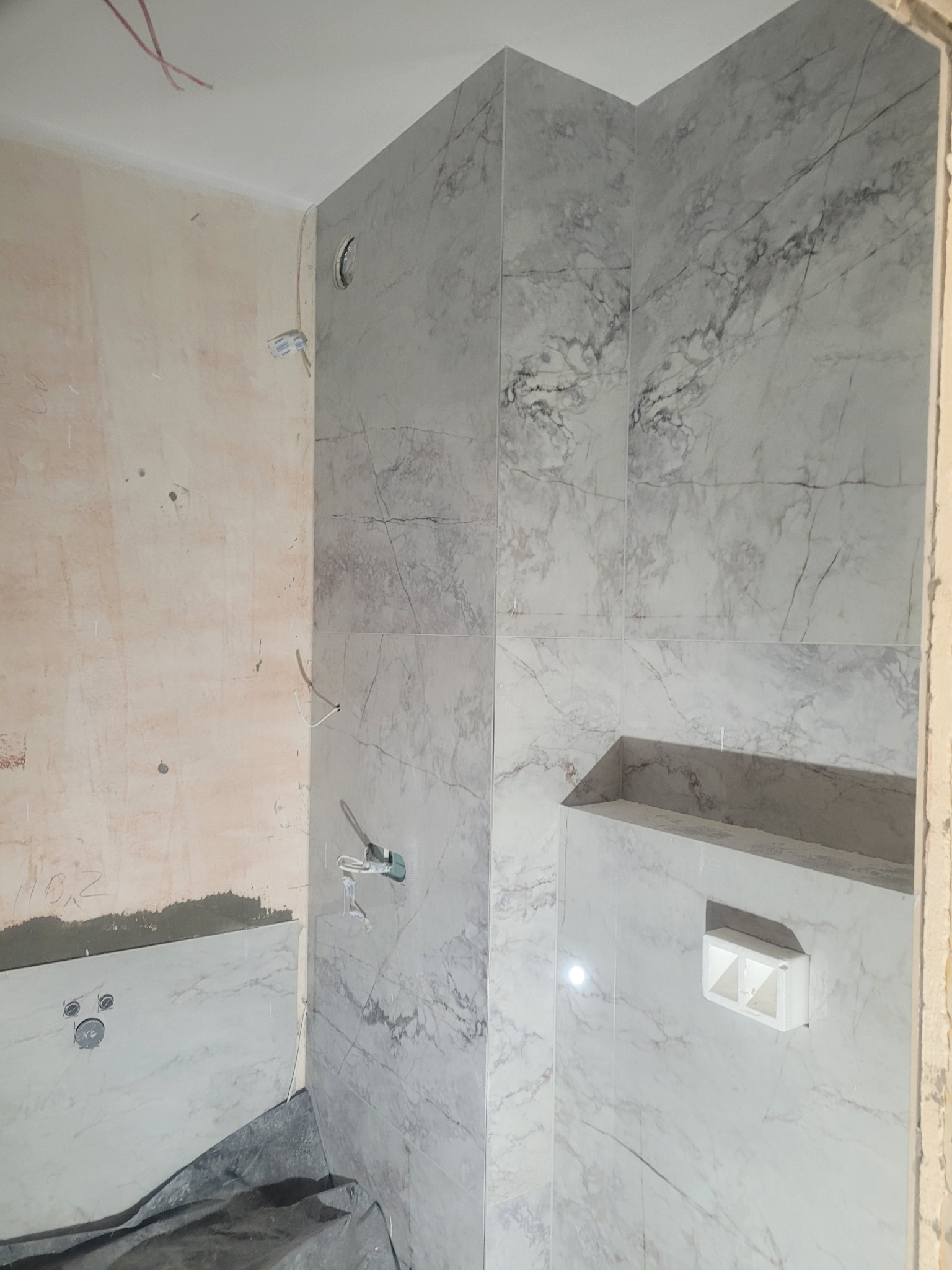 Łazienka w trakcie prac: ściana częściowo pokryta płytkami imitującymi marmur, widoczne wystające kable i rury, surowa ściana obok, wnęka z półką w zabudowie.