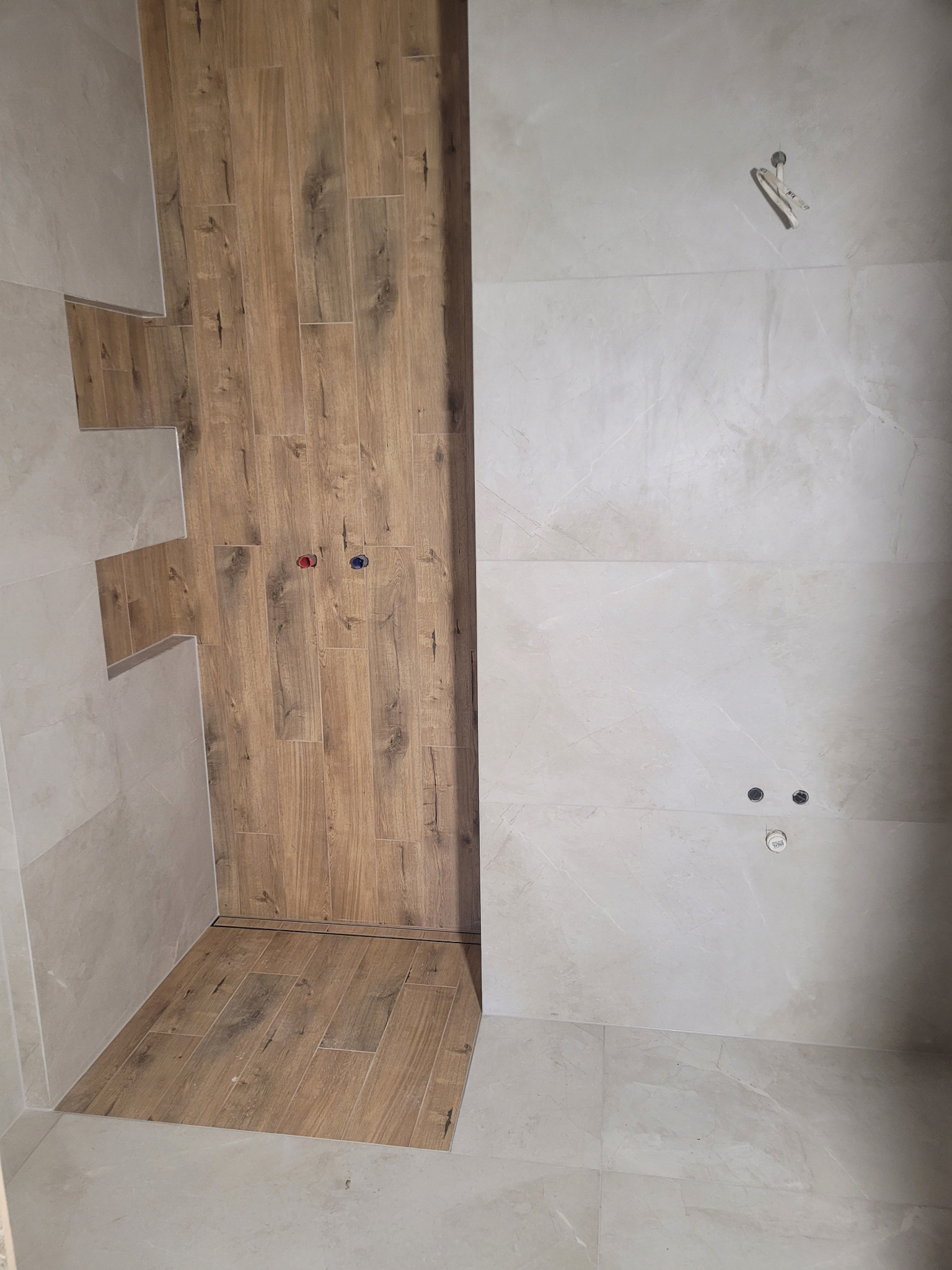 Wnętrze łazienki w trakcie prac wykończeniowych, z drewnopodobnymi płytkami na ścianie prysznica i jasnymi płytkami imitującymi marmur na pozostałych ścianach, widoczne otwory na armaturę.