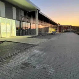 Szara kostka brukowa ułożona przed nowoczesnym budynkiem z przeszkloną fasadą, widok z perspektywy chodnika podczas zachodu słońca.