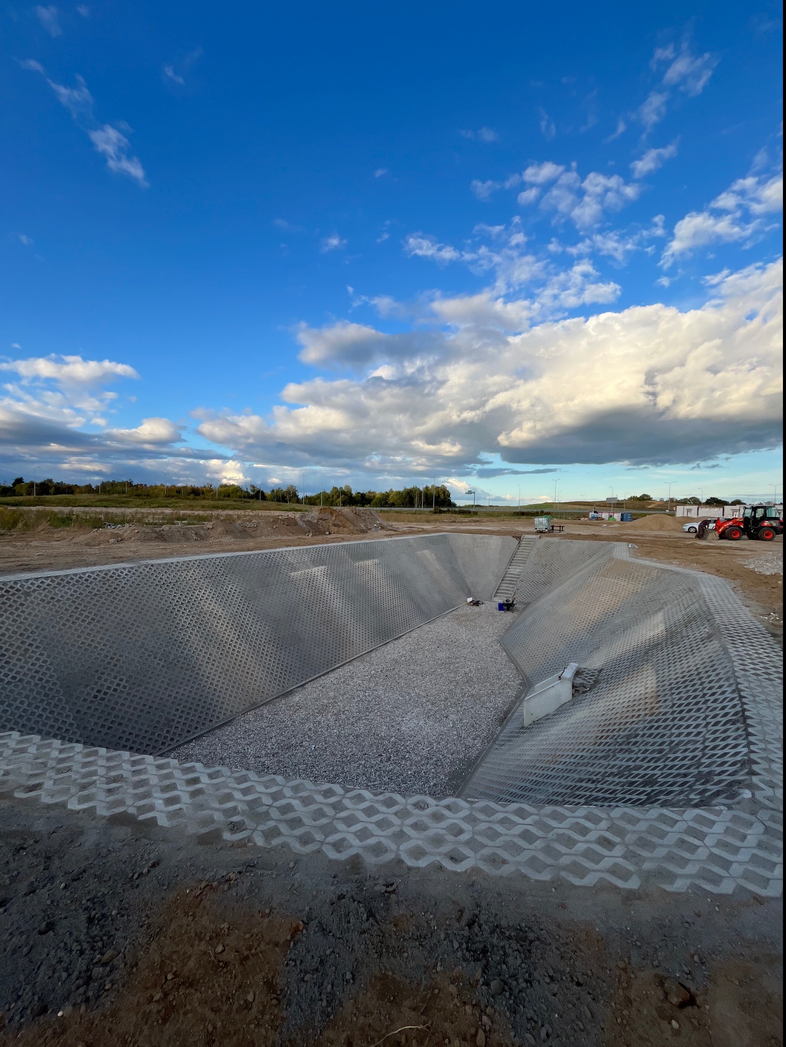 Skarpa zabezpieczona ażurowymi płytami betonowymi, wypełniona żwirem, widok z góry na teren budowy z błękitnym niebem i obłokami.