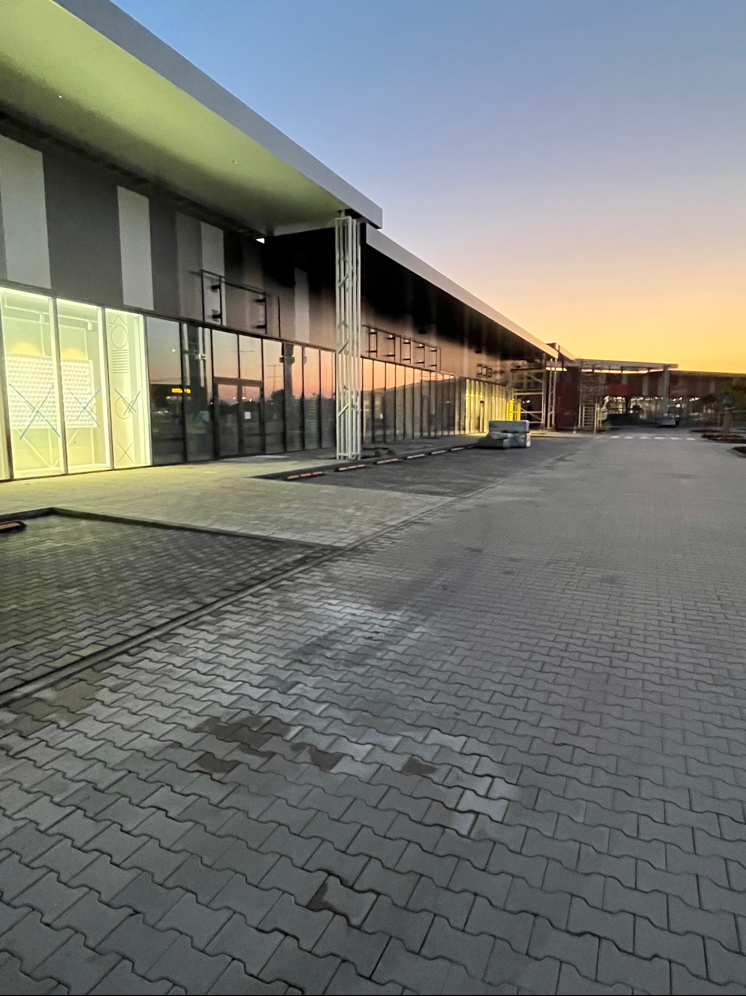 Szara kostka brukowa ułożona przed nowoczesnym budynkiem z przeszkloną fasadą, widok z perspektywy chodnika podczas zachodu słońca.