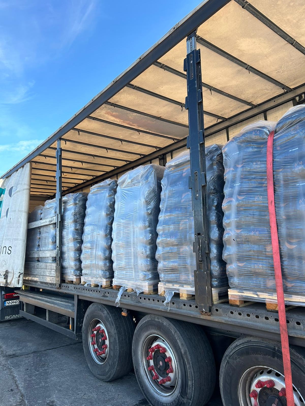 Ciężarówka z otwartą plandeką załadowana paletami z workami, owiniętymi folią stretch, zabezpieczonymi czerwoną taśmą transportową.