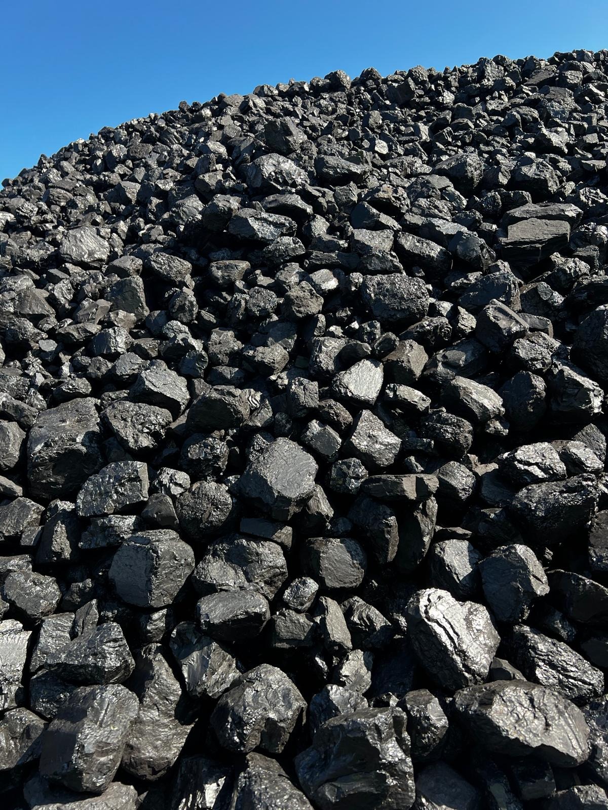 Stos czarnego węgla kamiennego pod błękitnym niebem, oświetlony z góry, z widocznymi błyszczącymi powierzchniami poszczególnych brył.