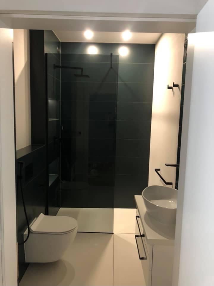 Nowoczesna łazienka z czarnymi płytkami na ścianach kabiny prysznicowej, białą umywalką nablatową i podwieszaną toaletą, oświetlona punktowymi lampami sufitowymi.