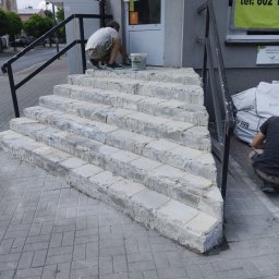 KM.BuD - Pracownik maluje wejście do budynku z kamiennymi schodami, widoczny kubeł z farbą i reklama na budynku w tle.