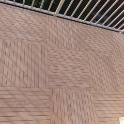 KM.BuD - Wykończony balkon z drewnopodobnych, kwadratowych paneli ułożonych w szachownicę, widoczne białe barierki.