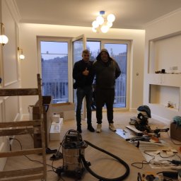 KM.BuD - Dwaj mężczyźni na tle jasnego, odnowionego wnętrza z parkietem, widoczne narzędzia budowlane, drabina malarska i odkurzacz przemysłowy, światło lamp sufitowych i kinkietów.