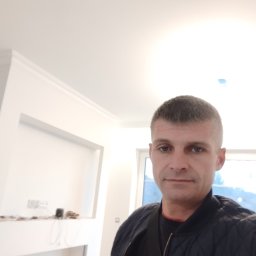 KM.BuD - Mężczyzna w kurtce na tle białego wnętrza w trakcie wykańczania, widoczne listwy przysufitowe i przygotowane wnęki na ścianie.