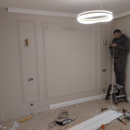 KM.BuD - Instalacja listew dekoracyjnych na ścianie z ozdobnymi panelami; pracownik na drabinie, widoczne narzędzia i materiały na parkiecie w jodełkę, nowoczesna lampa sufitowa.