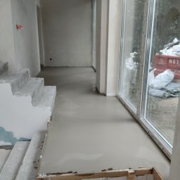 KM.BuD - Wylewka samopoziomująca na podłodze w surowym wnętrzu, widoczne schody w stanie deweloperskim i okno z widokiem na zewnątrz, w tle kontener na gruz.