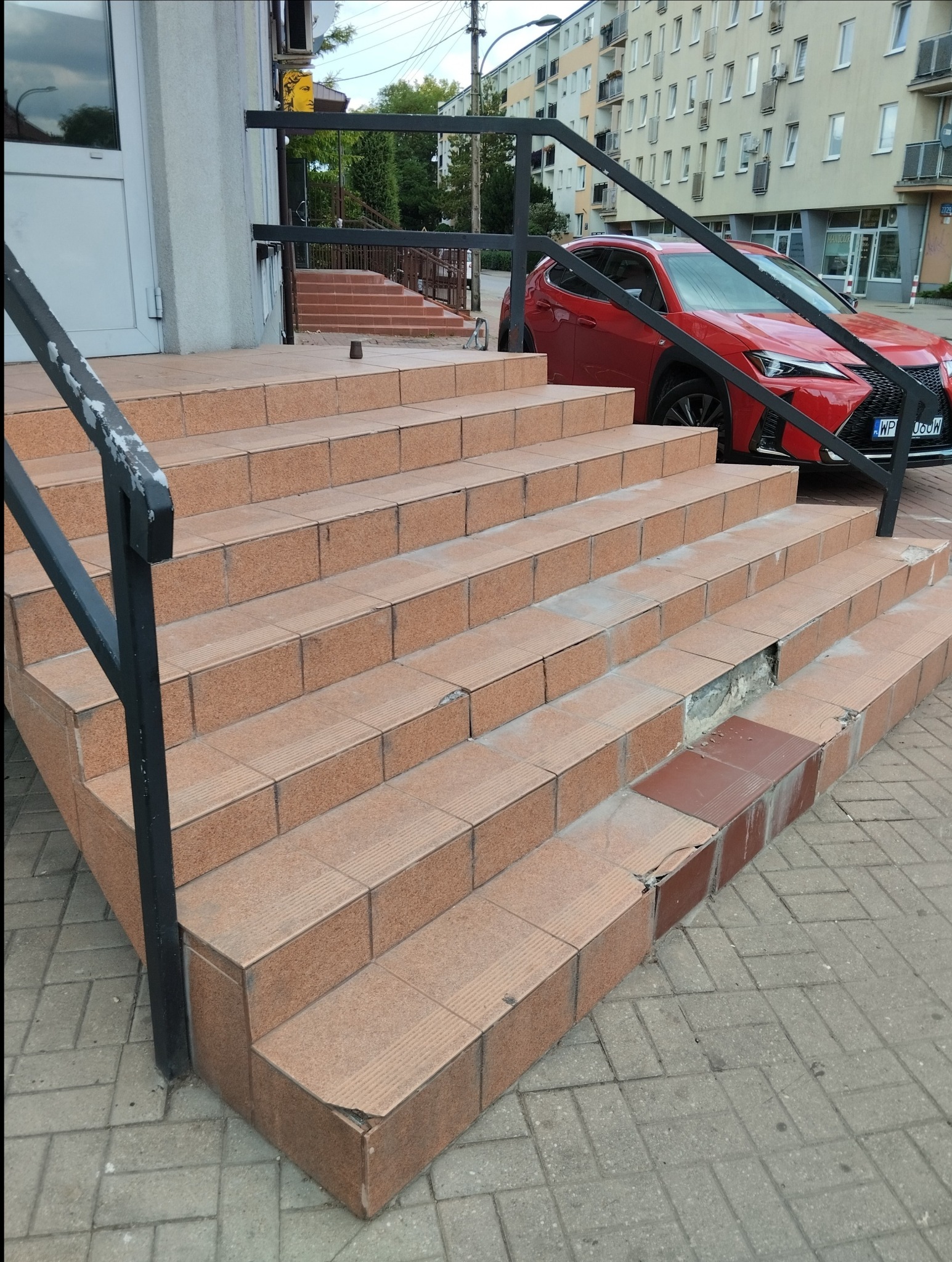 Zewnętrzne schody wejściowe z uszkodzonymi płytkami, widoczny ubytek i brakujące fragmenty, na tle budynku mieszkalnego i czerwonego samochodu.