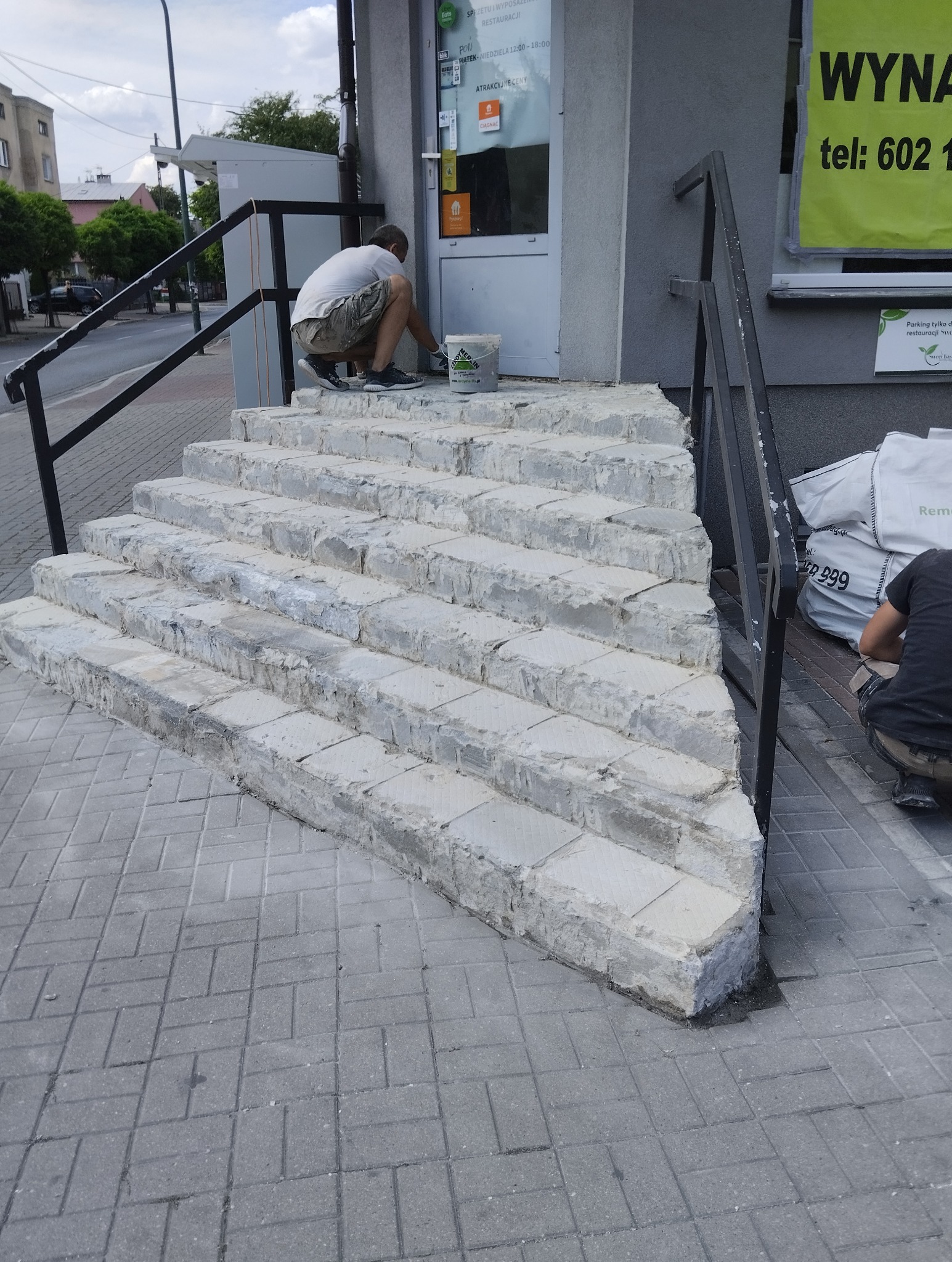 Pracownik maluje wejście do budynku z kamiennymi schodami, widoczny kubeł z farbą i reklama na budynku w tle.