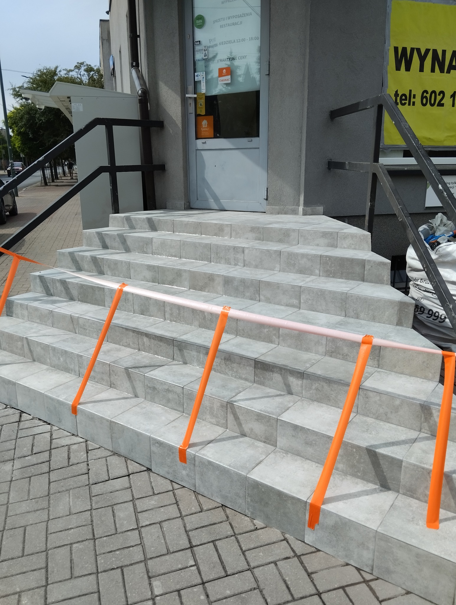 Wykończone schody wejściowe do budynku z szarych płytek, zabezpieczone pomarańczową taśmą ostrzegawczą, widok z dołu na zewnątrz.