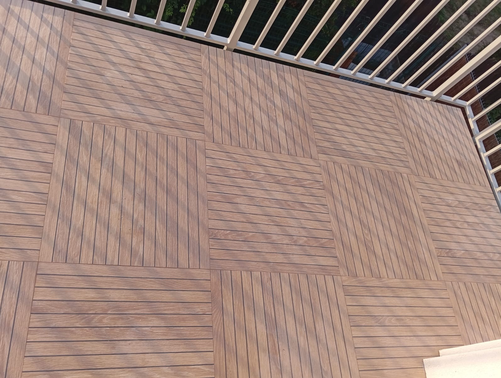 Wykończony balkon z drewnopodobnych, kwadratowych paneli ułożonych w szachownicę, widoczne białe barierki.