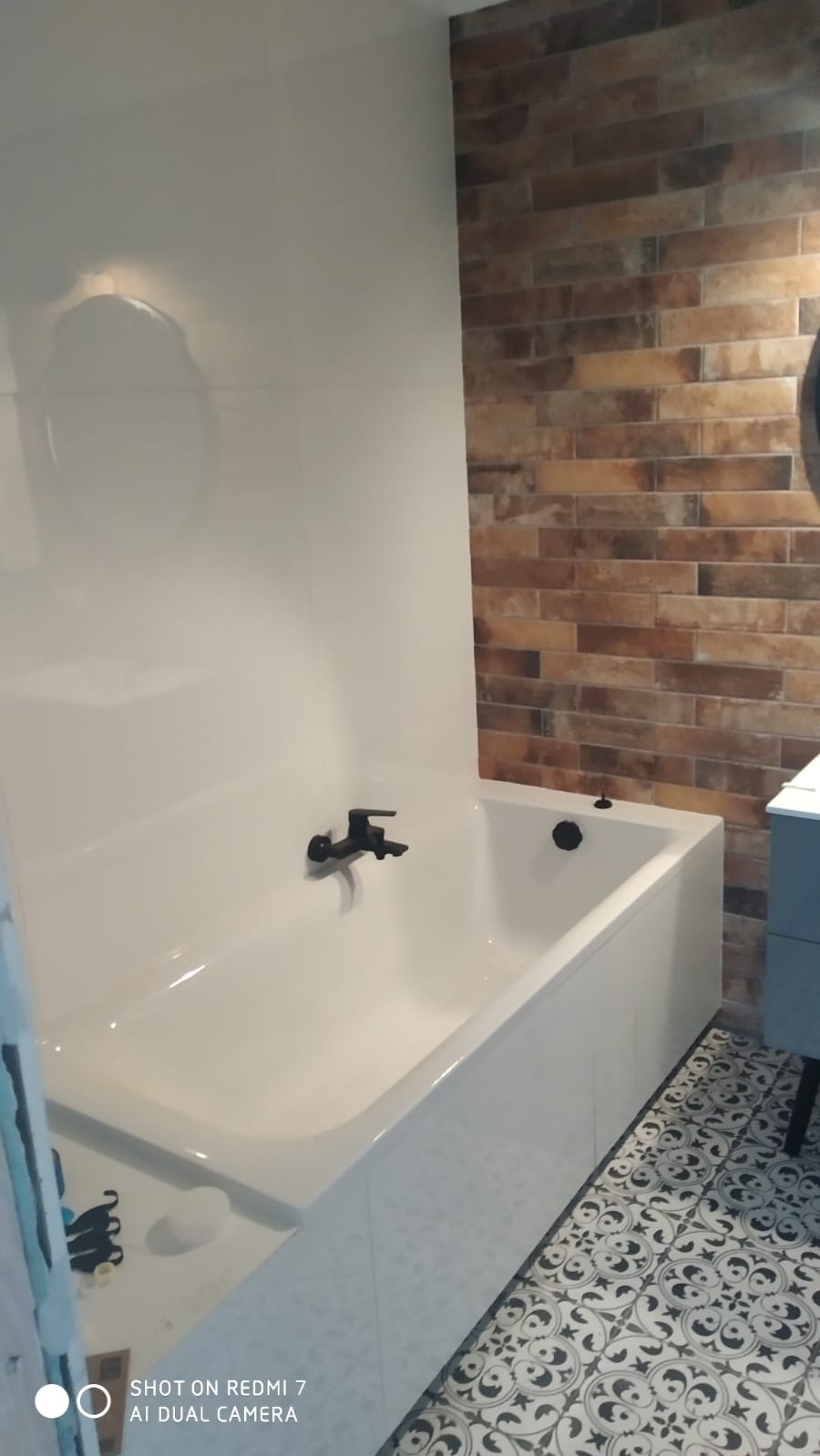 Biała wanna z czarną armaturą na tle ściany z cegieł i wzorzystej podłogi w łazience po remoncie.