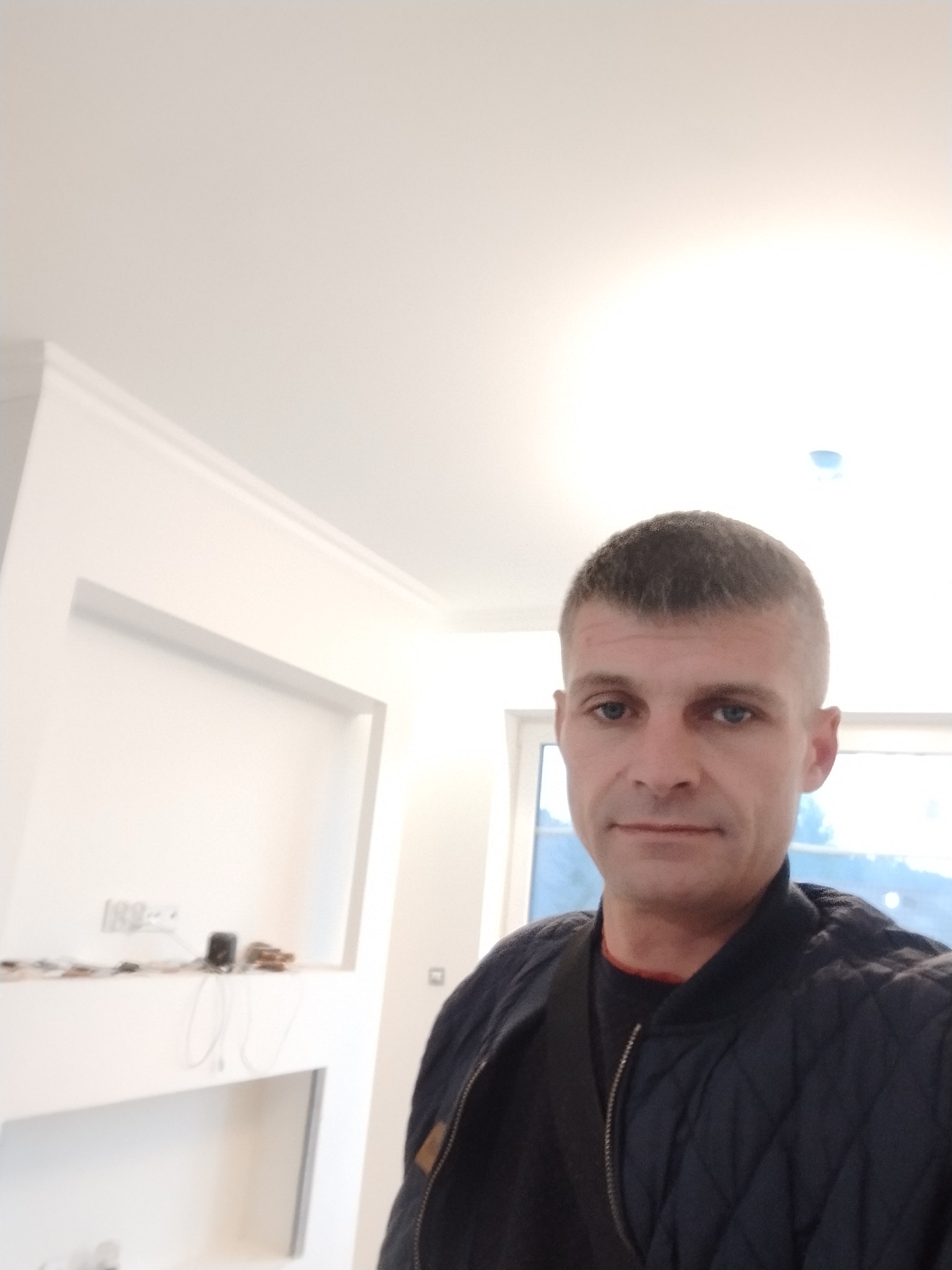 Mężczyzna w kurtce na tle białego wnętrza w trakcie wykańczania, widoczne listwy przysufitowe i przygotowane wnęki na ścianie.